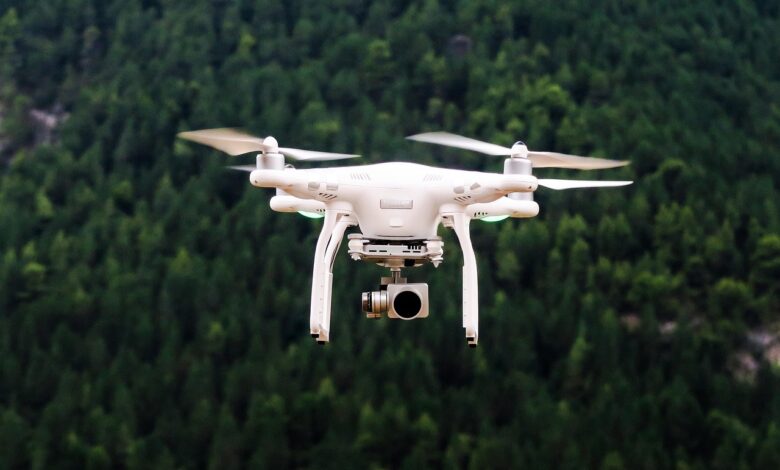 White Quadcopter Drone Hovering in Front of a Forested Background With Camera Attached