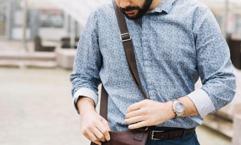 A Man is Looking at a Mini Bag for Men—a Compact Crossbody Leather Mini Bag With a Flap—held in Front of Him, Showing Its Detail
